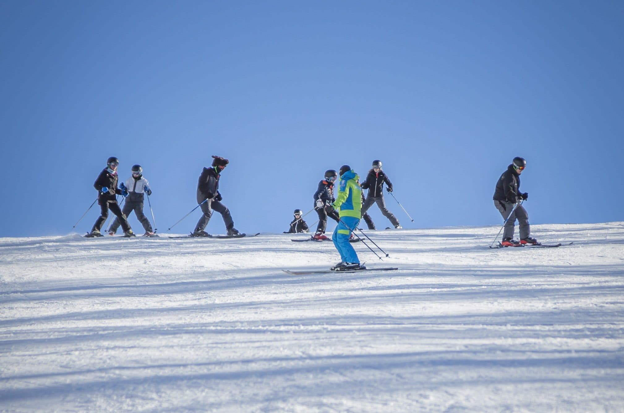 A ski instructor in a bright jacket leads a group of kids and adults down a gentle groomed slope