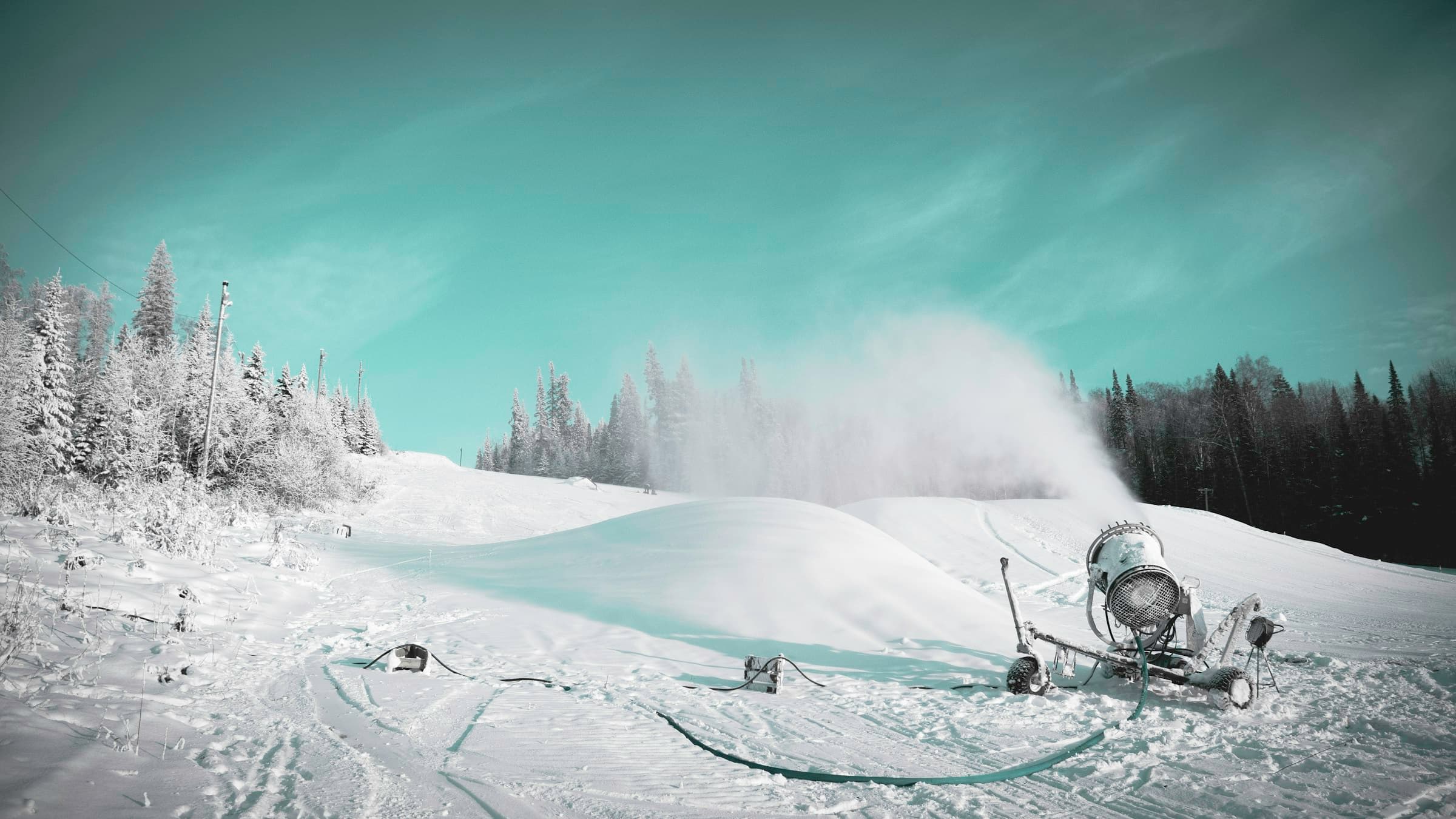 A snowmaking gun blowing a plume of man-made snow across a groomed ski slope lined with frost-covered evergreens